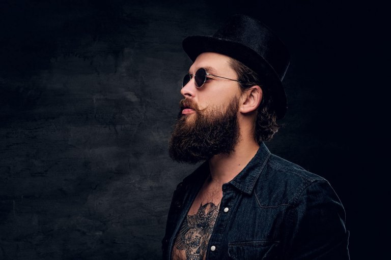 Groomed bearded man with tattooed chest is posing for photographer at dark photo studio.
